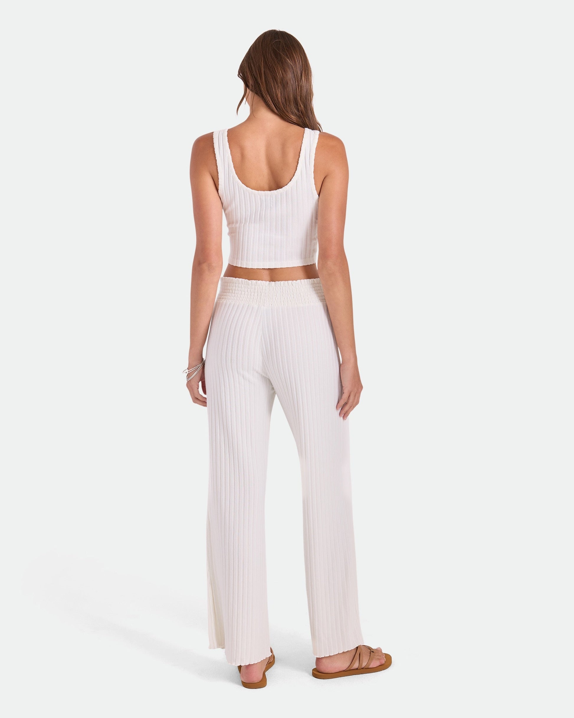 A woman with long brown hair faces away, wearing Volcom Women's Sun Lounge Pants—relaxed fit with elastic waistband—and a white ribbed crop top and sandals, shown on a plain white background.