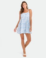 A woman in the Volcom Women's Beachmoonstone dress and beige sandals stands against a white background, smiling gently with one hand by her side and the other slightly raised.