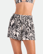 A woman wears Volcom Women's Funtastical Viscose Shorts in a black ecru tropical leaf print with an elastic waistband, paired with a matching bikini top against a plain light-colored background.