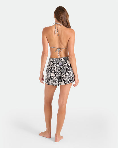 A woman faces away, barefoot against a white background, wearing Volcom Women's Funtastical Viscose Shorts with an elastic waistband in a Black Ecru print and a halter-neck striped bikini top.