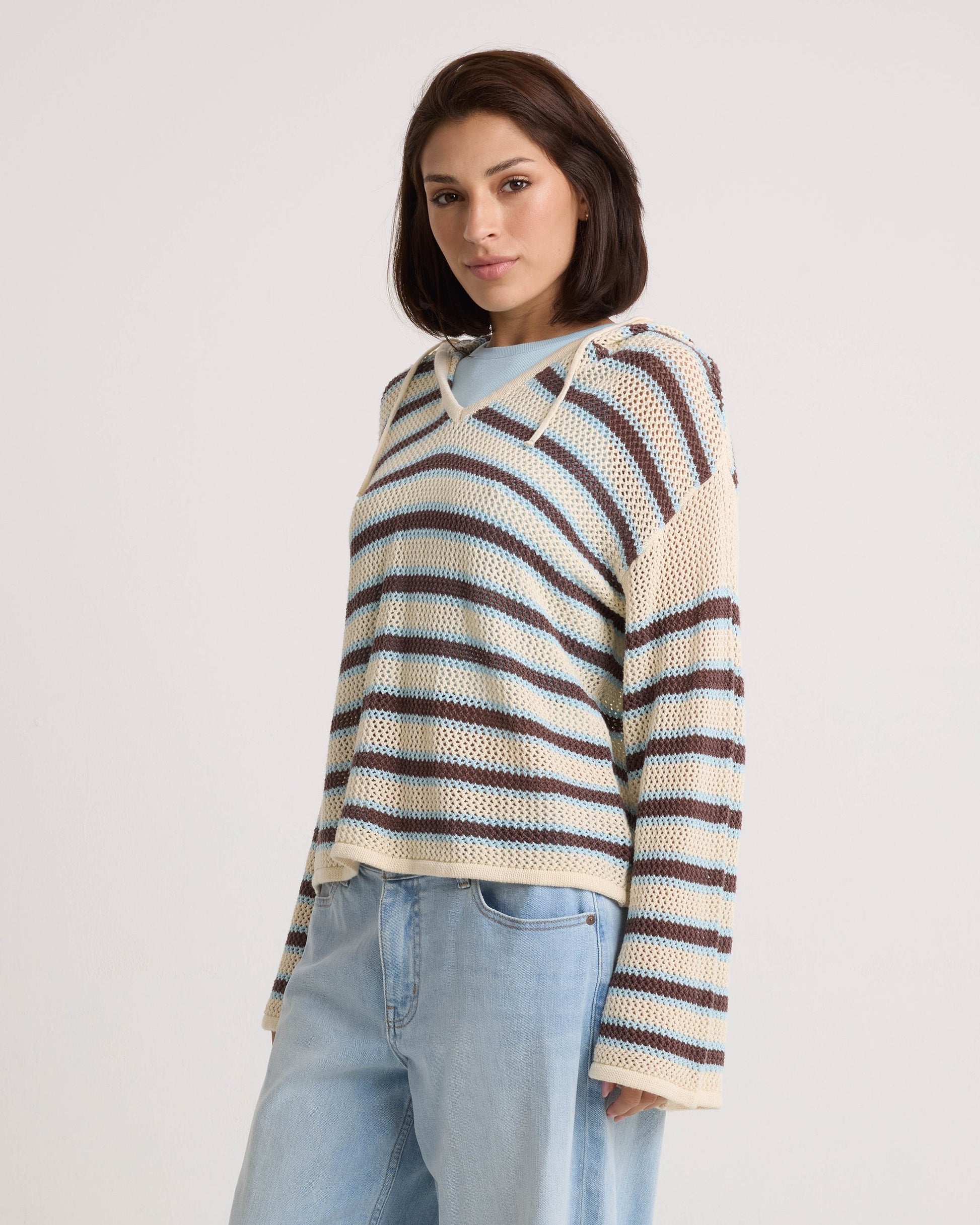 A woman with short brown hair models the Volcom Women's Bonfire Hoodie in beige, blue, and brown stripes with light blue jeans. She stands against a plain white background, looking confidently at the camera.