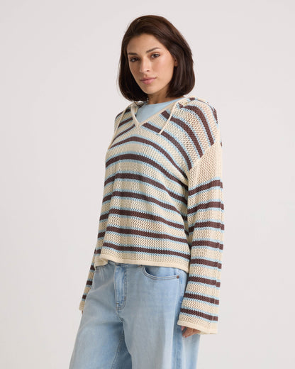A woman with short brown hair models the Volcom Women's Bonfire Hoodie in beige, blue, and brown stripes with light blue jeans. She stands against a plain white background, looking confidently at the camera.