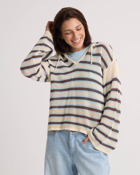 A smiling woman with short brown hair models the Volcom Women's Bonfire Hoodie—a loose, striped cotton sweater with a crochet texture—paired with light blue jeans. She poses against a plain background, hand in pocket and touching her head.