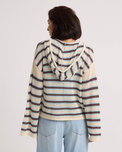 A woman with short dark hair faces away from the camera in a Volcom Women's Bonfire Hoodie—a hooded, crochet-textured cotton pullover with blue, brown, and cream stripes—paired with light blue jeans against a plain white background.