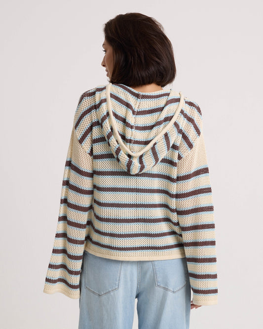 A woman with short dark hair faces away from the camera in a Volcom Women's Bonfire Hoodie—a hooded, crochet-textured cotton pullover with blue, brown, and cream stripes—paired with light blue jeans against a plain white background.