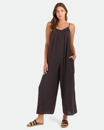A woman with long hair wears the Volcom Women's Ruffle Up Romper, a loose, sleeveless, dark romper with spaghetti straps and wide legs. She stands with one hand in her pocket and wears black sandals against a plain white background.