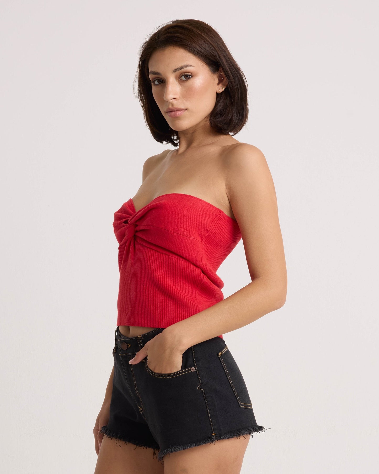 A woman with straight brown hair stands sideways against a white background, wearing the Volcom Women's Merlin Twist Top in persimmon and black frayed-hem denim shorts.