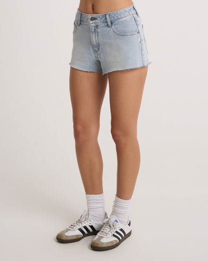 A person wears Volcom Women's Cruz Control light blue chambray wash denim shorts, white socks, and white Adidas sneakers with black stripes, standing against a plain white background. Only the lower half of their body is visible.
