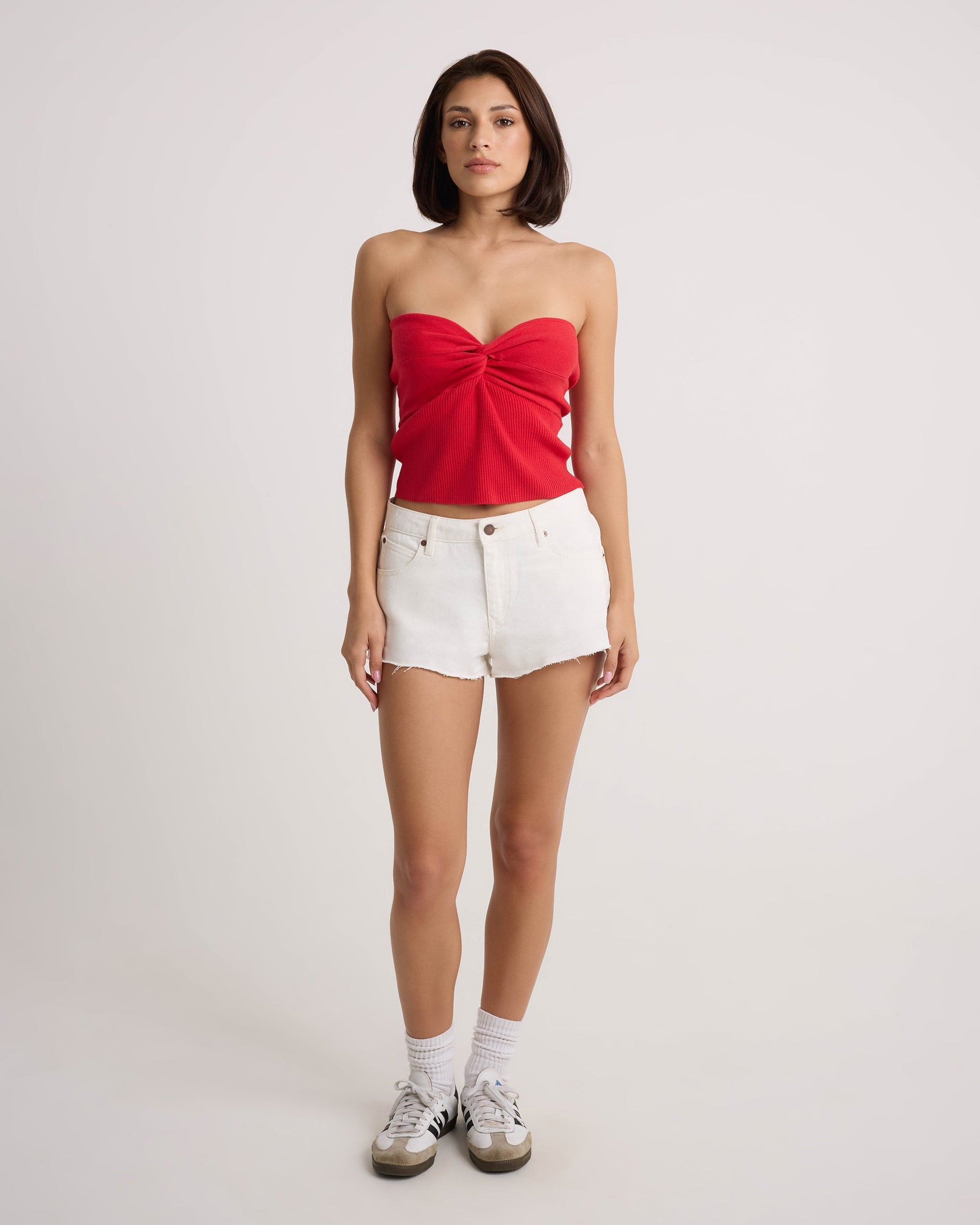 A woman faces forward wearing the Volcom Women's Cruz Control red strapless top and vintage white high-rise denim shorts, styled with white socks and sneakers, against a plain light background.