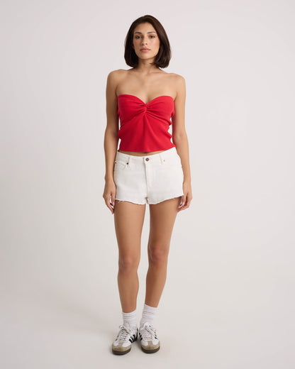 A woman faces forward wearing the Volcom Women's Cruz Control red strapless top and vintage white high-rise denim shorts, styled with white socks and sneakers, against a plain light background.