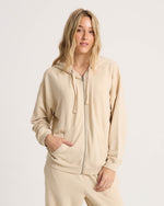 A woman with long blonde hair wears the Volcom Women's Lil Frenchie Zip Hoodie, standing against a plain white background, facing forward with one hand in her pocket.