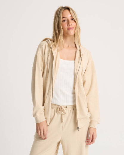 A woman with long blonde hair wears the Volcom Women's Lil Frenchie Zip Hoodie in beige and matching drawstring pants over a white ribbed tank top, standing against a plain light background and smiling at the camera.