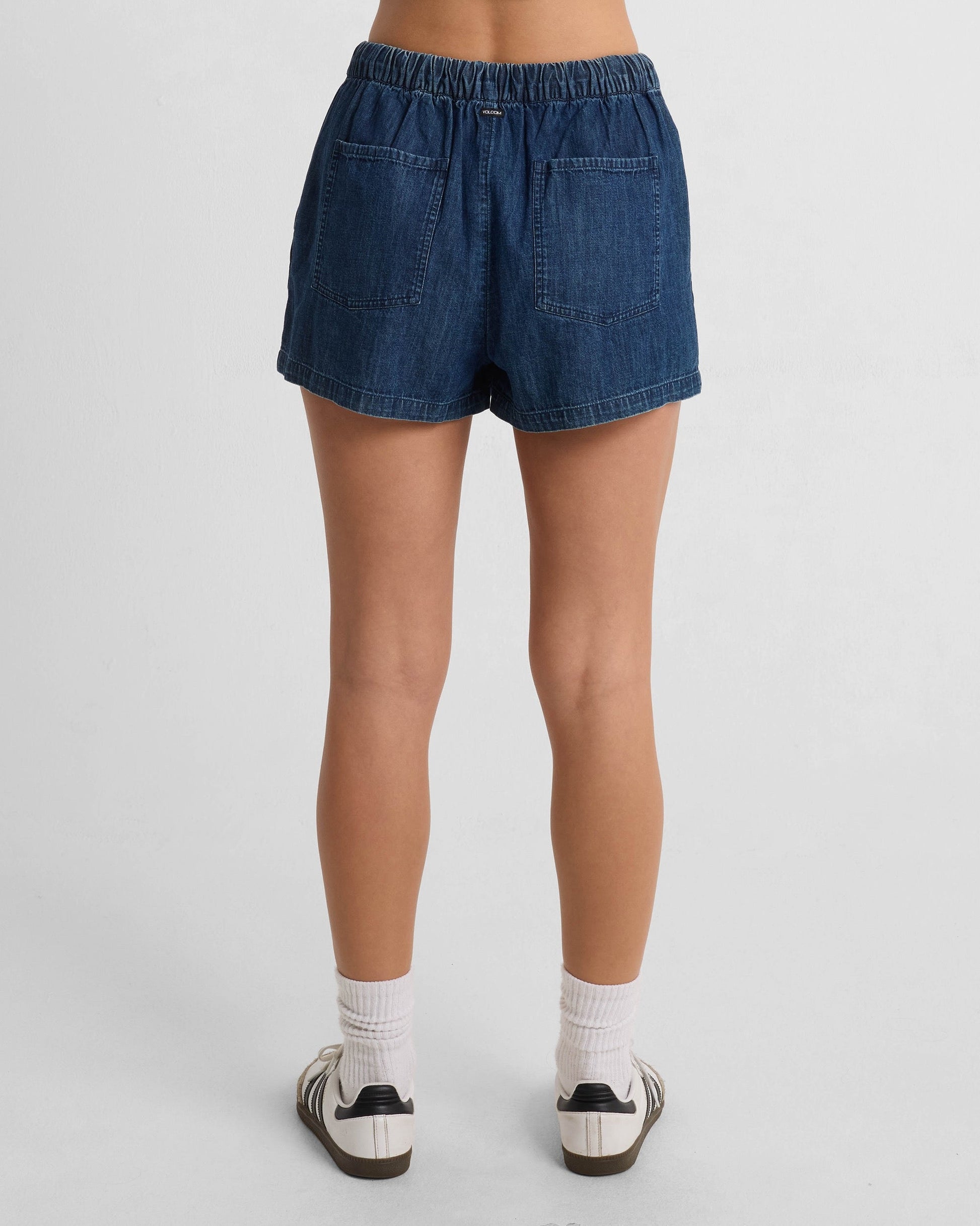 A person is standing facing away, wearing Volcom Women's Stone Shine Shorts made from 100% cotton with two back pockets, paired with white socks and white sneakers with black stripes, against a plain white background.