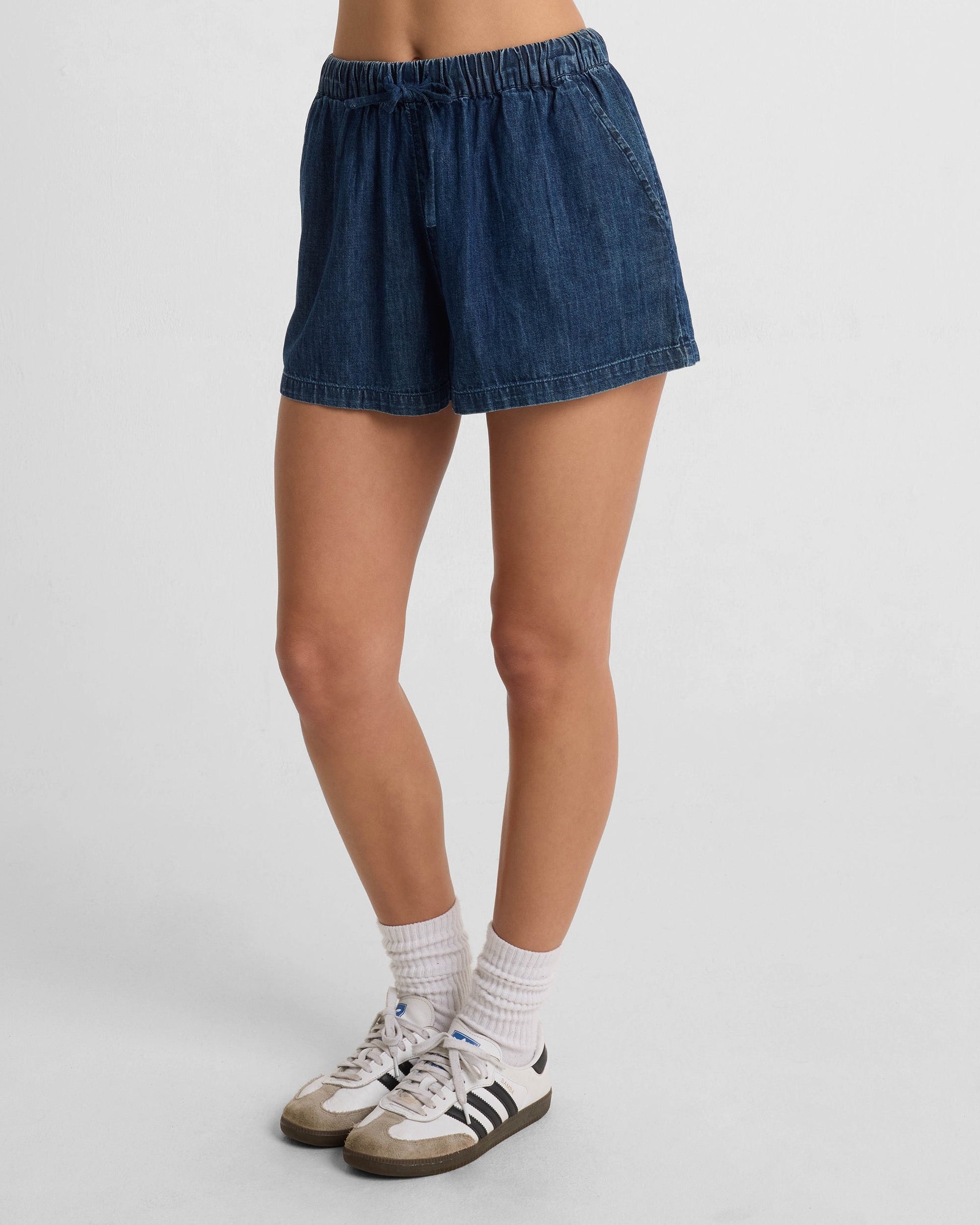 A person wears Volcom Women's Stone Shine Shorts with white socks and white sneakers featuring black stripes, standing against a plain white background.