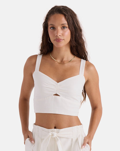 A woman with long wavy brown hair wears the Volcom Women's Bow Chicka Tank, a vintage white crop top featuring a front knot detail, paired with matching high-waisted pants. She stands with hands in pockets against a plain white background.