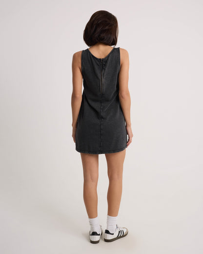 A woman with shoulder-length dark hair faces away, wearing the Volcom Women's Glam Crop Dress in Vintage Black with a visible back zipper, paired with white socks and black-and-white sneakers against a plain background.