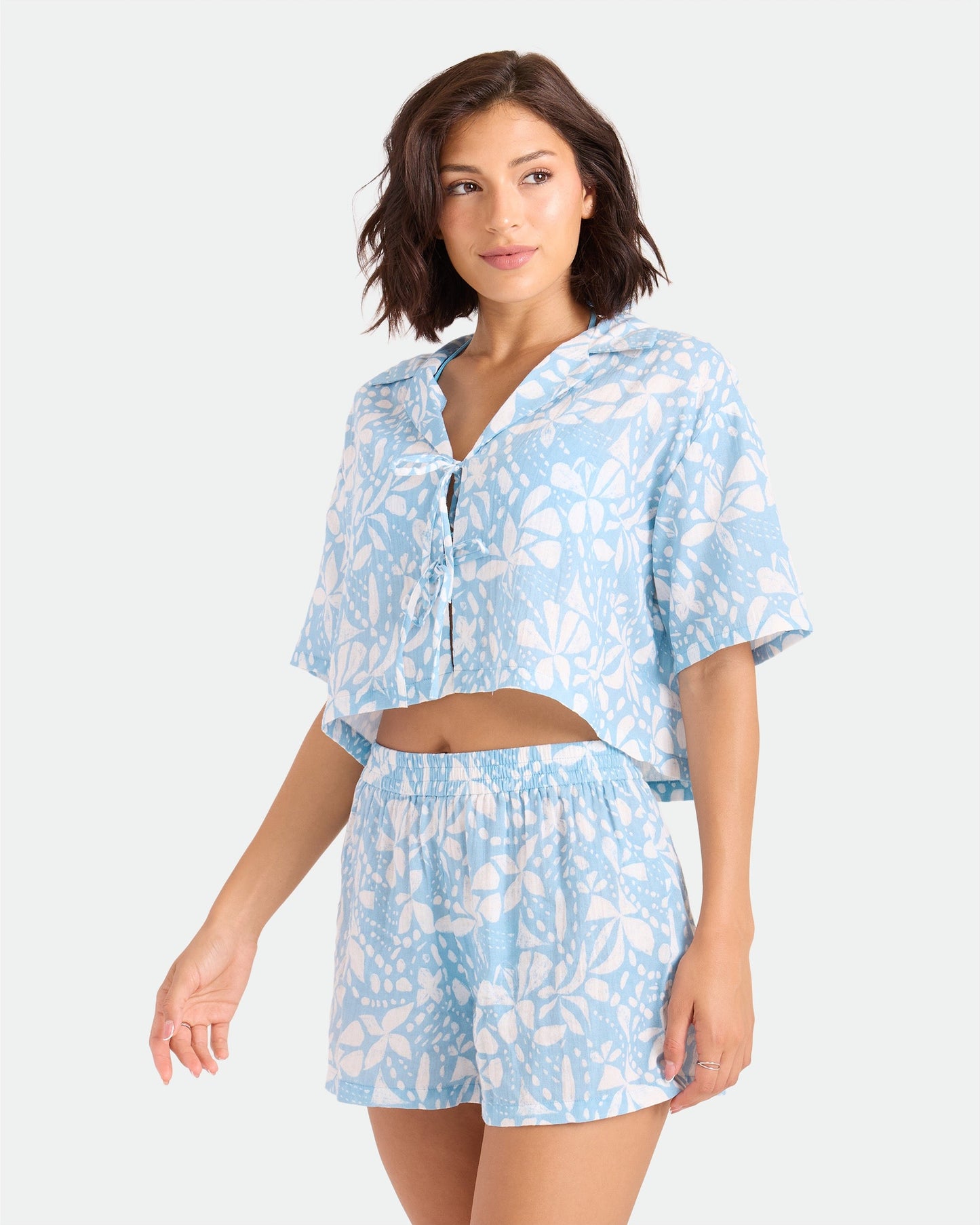 A woman wears Volcom's Women's Funtastical Gauze Shorts—high-waisted, elasticated, double-gauze cotton with a white floral print—paired with a matching light blue crop top, standing against a plain light background.