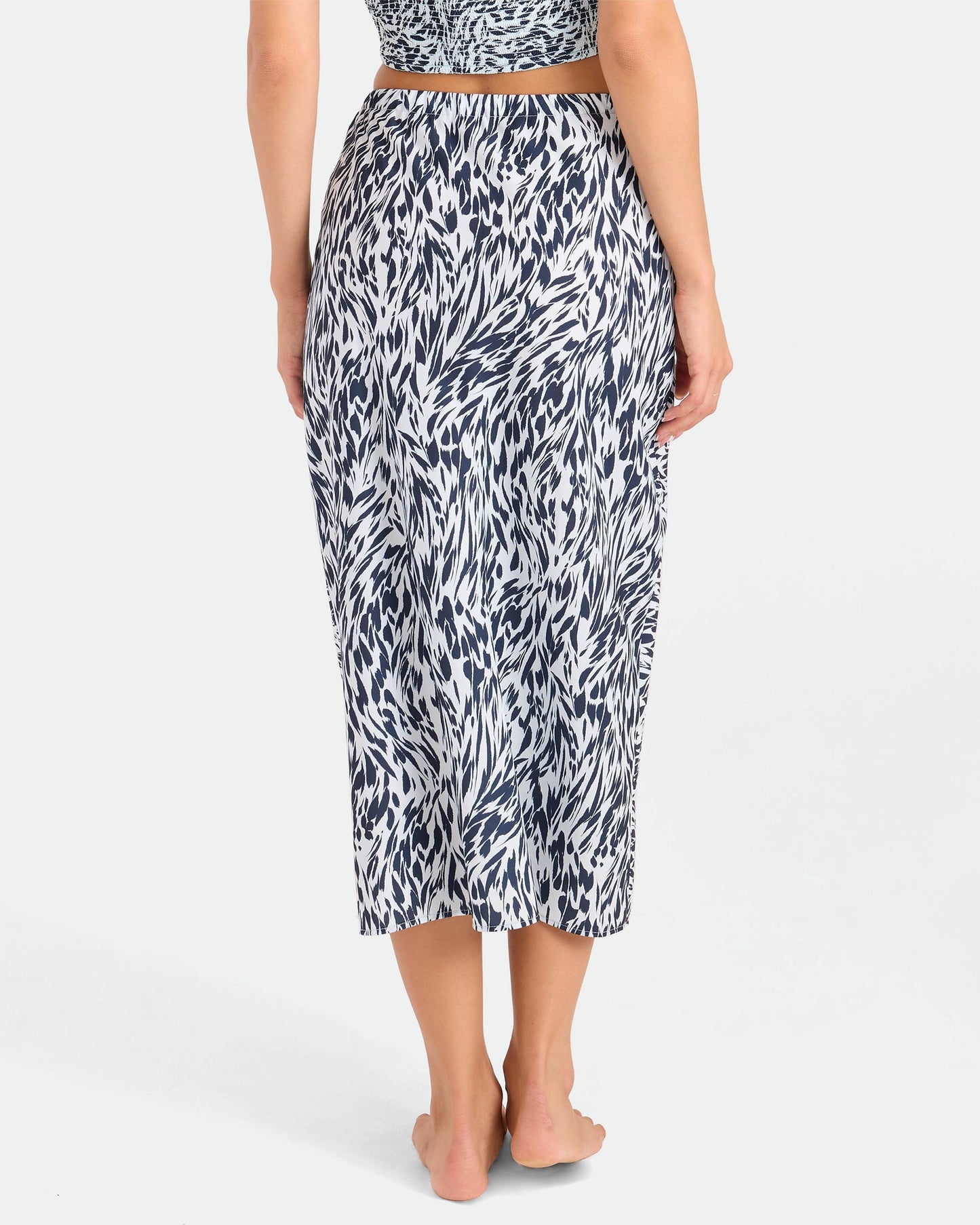 A person stands barefoot, facing away, wearing the Volcom Women's Coco Ho Sandy Parts Skirt—a high-waisted midi skirt with a black and white abstract pattern. The background is plain and light-colored.