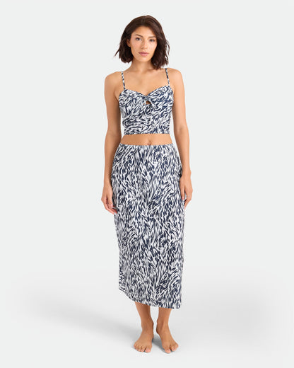 A woman stands barefoot in the Volcom Women's Coco Ho Sandy Parts Skirt set—featuring a fitted crop top and high-waisted, calf-length skirt with a blue and white abstract pattern—flowing elegantly against a plain white background.