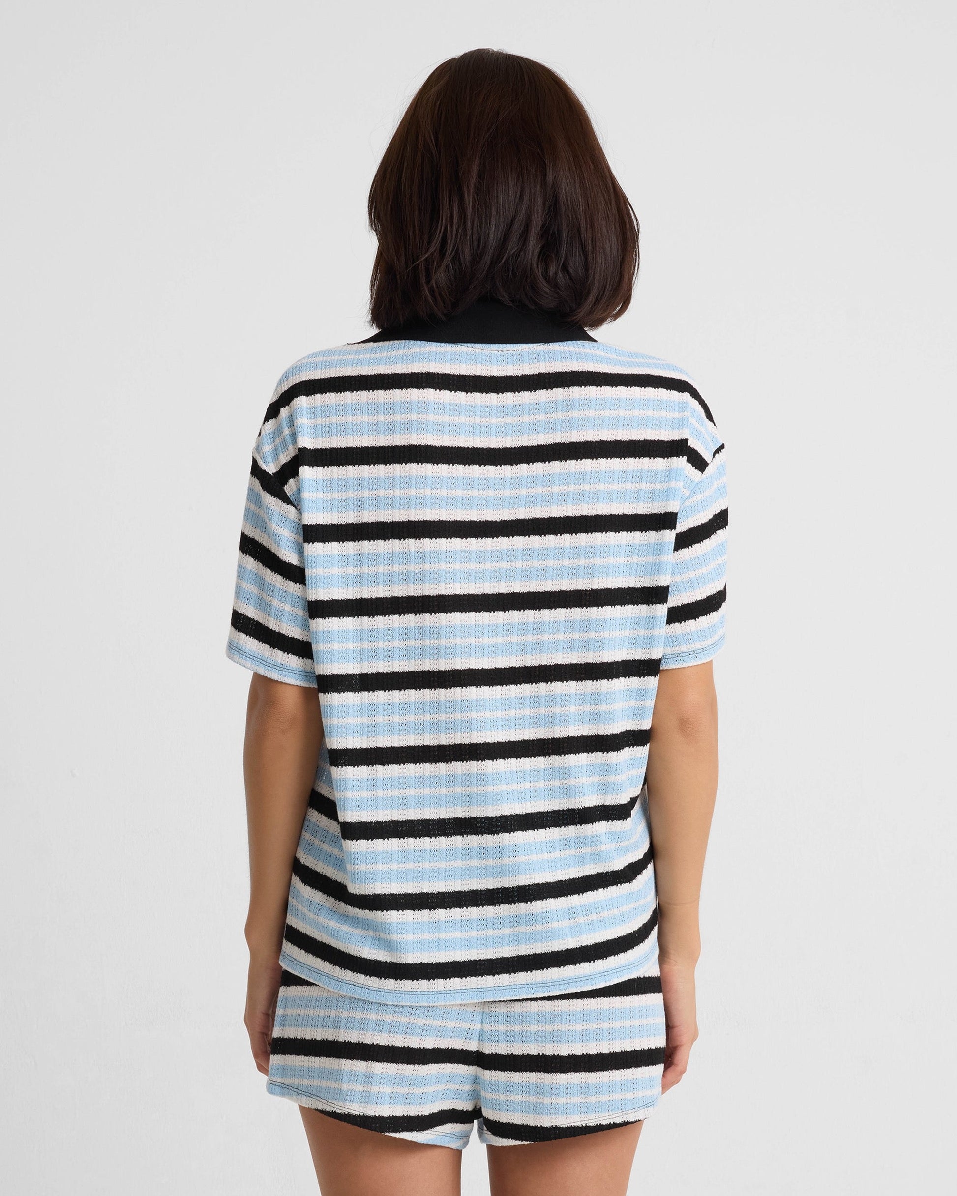 A person with shoulder-length dark hair stands with their back to the camera, wearing Volcom's Women's Coco Ho Crochet Shorts in light blue, white, and black stripes. The set has relaxed fit short sleeves. The background is plain white.