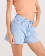 A person wearing Volcom Women's Stoned Bf Short in Blue Bird light wash (5-inch inseam, boyfriend fit) and a peach cropped tee, one hand in pocket. Only the midsection and upper legs show against a plain light background.