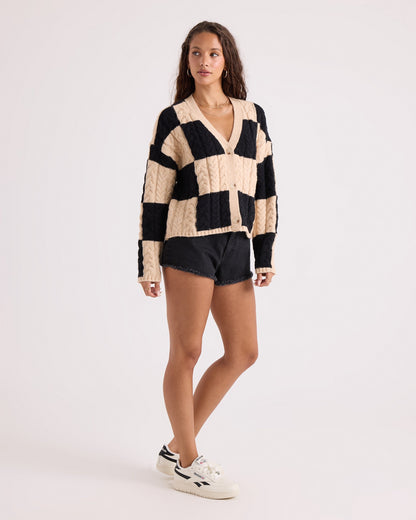 A woman with long wavy hair models the Volcom Women's Check You Sweater—a beige and black checkerboard patterned V-neck cardigan with a comfy knit and button front—paired with black denim shorts and white sneakers, standing against a plain white background.