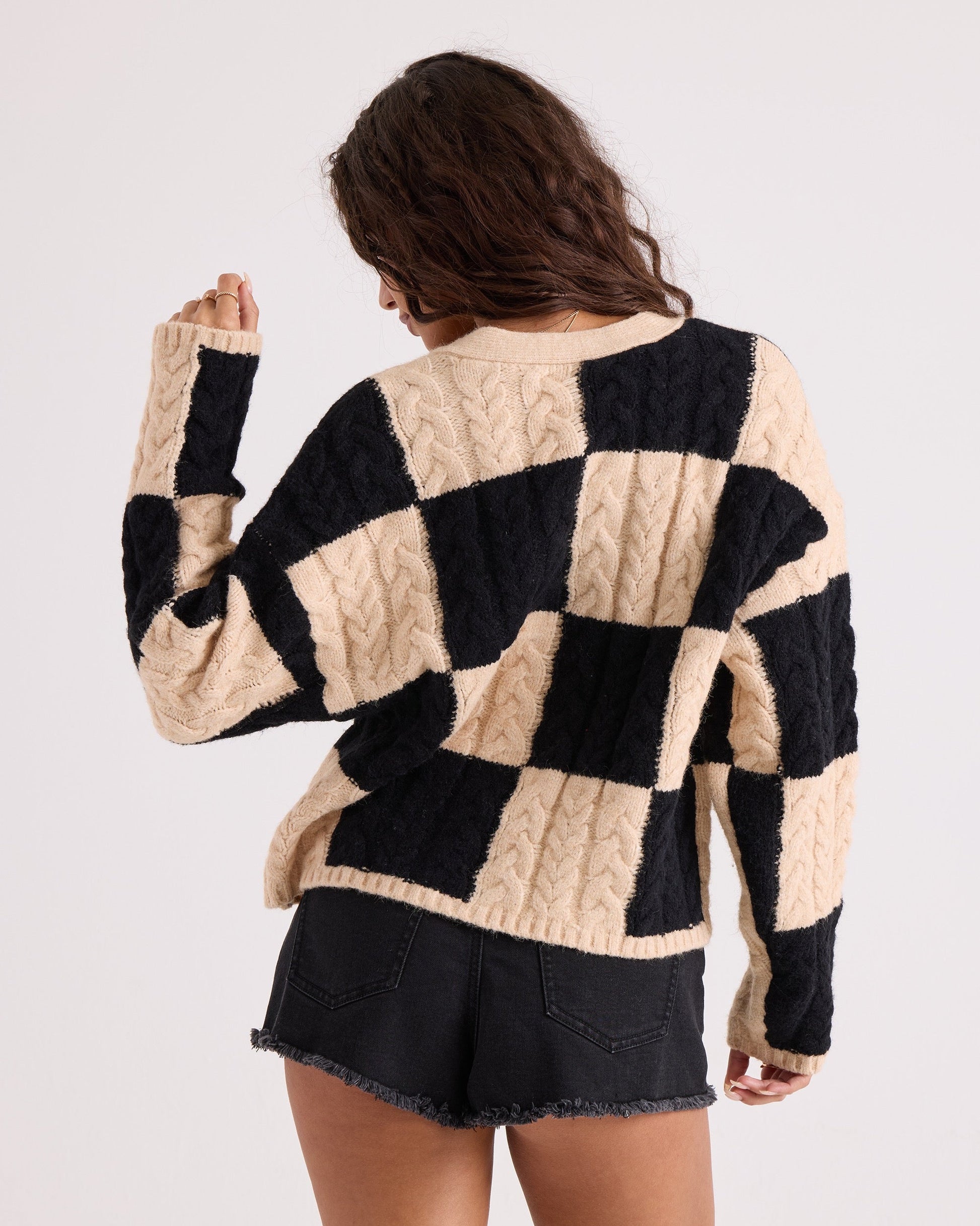 A person with long wavy hair, seen from behind, wears the Volcom Women's Check You Sweater—a comfy knit black and beige checkered cable-knit V-neck—paired with black denim shorts against a plain light background.