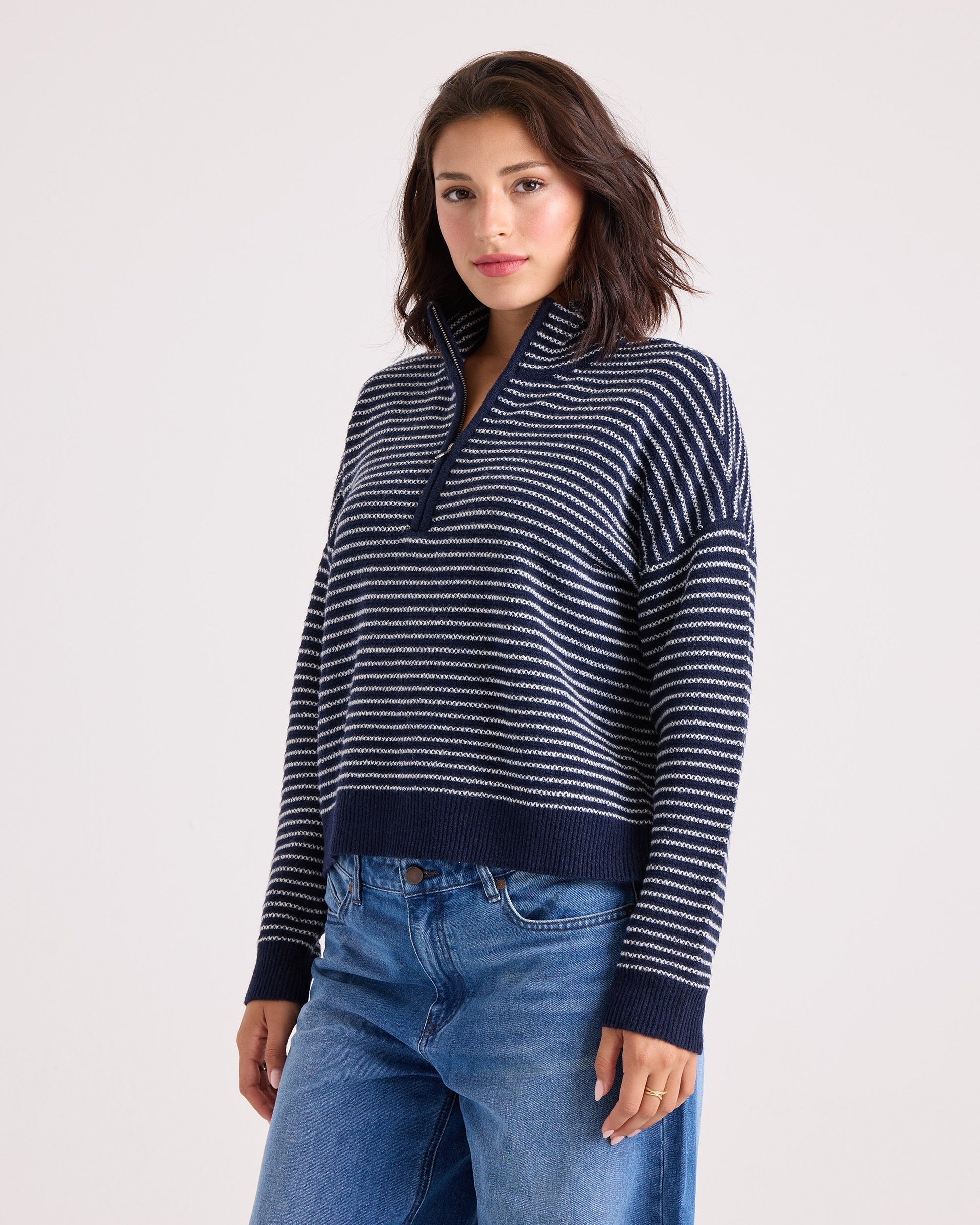 A woman wearing the Volcom Women's Sun Of Sand Sweater—a navy and white striped quarter-zip sweater with a collar—paired with blue jeans, stands against a plain light background, looking at the camera with a neutral expression.