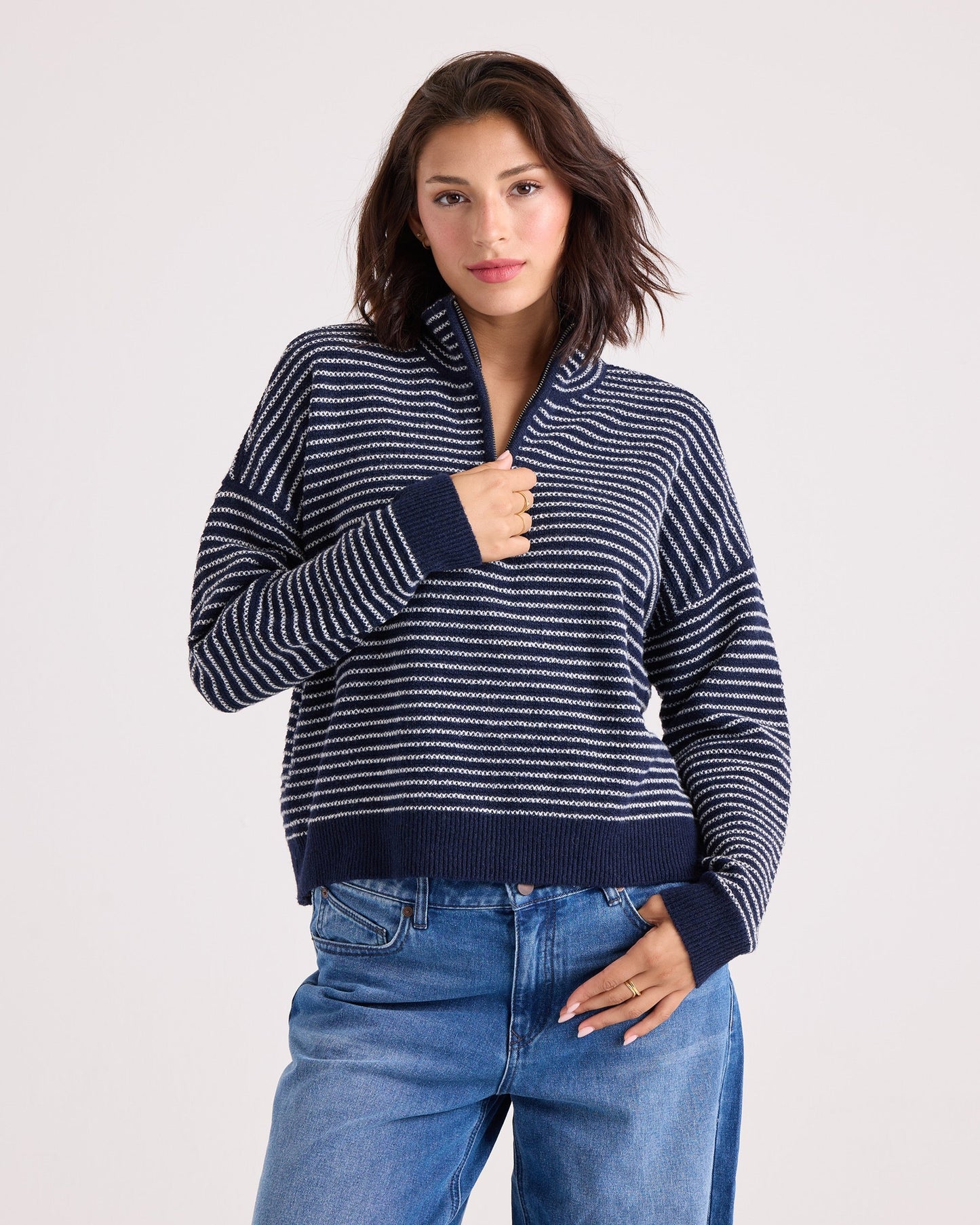 A woman with shoulder-length dark hair wears the Volcom Women's Sun Of Sand Sweater—a navy and white striped quarter-zip design—paired with blue jeans, standing against a plain light background with one hand touching her collar.