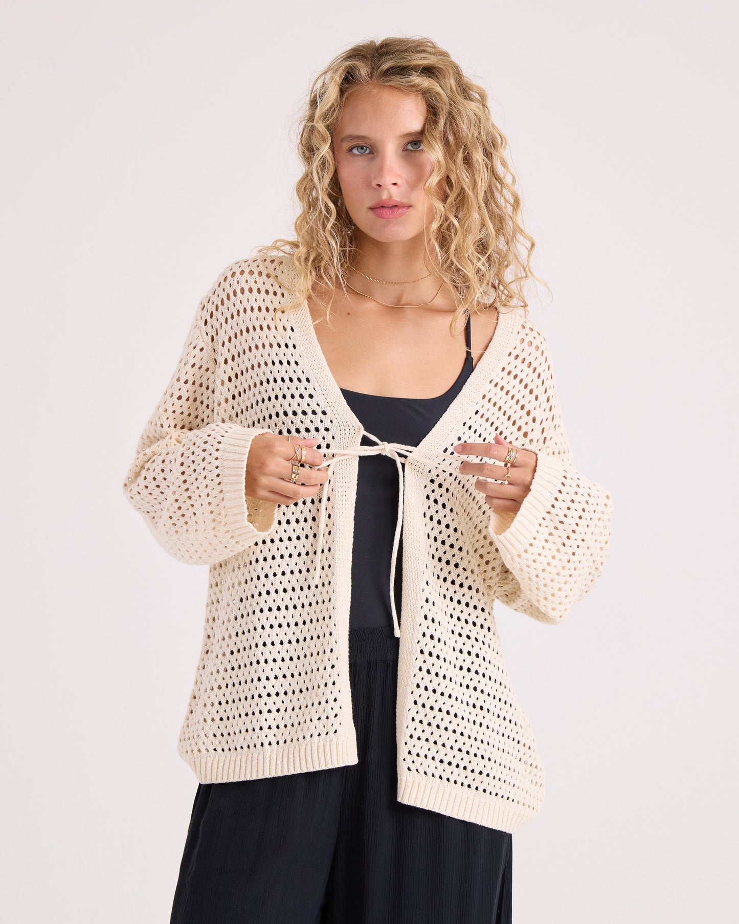 A woman with curly blonde hair wears the Volcom Women's Go Go Wrap, a cream-colored, open-knit tie-front cardigan, over a black top and pants, standing against a plain background.