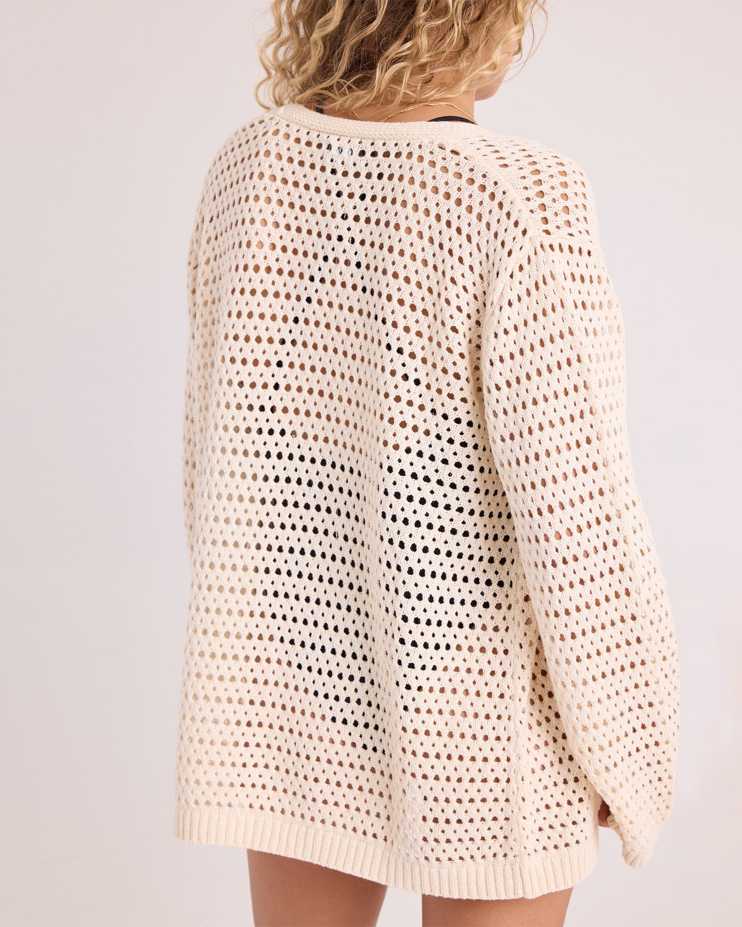 A person with curly blonde hair is shown from the back, standing against a plain, light background and wearing the Volcom Women's Go Go Wrap—a loose, cream-colored, open-knit sweater top crafted from a soft cotton-acrylic blend.