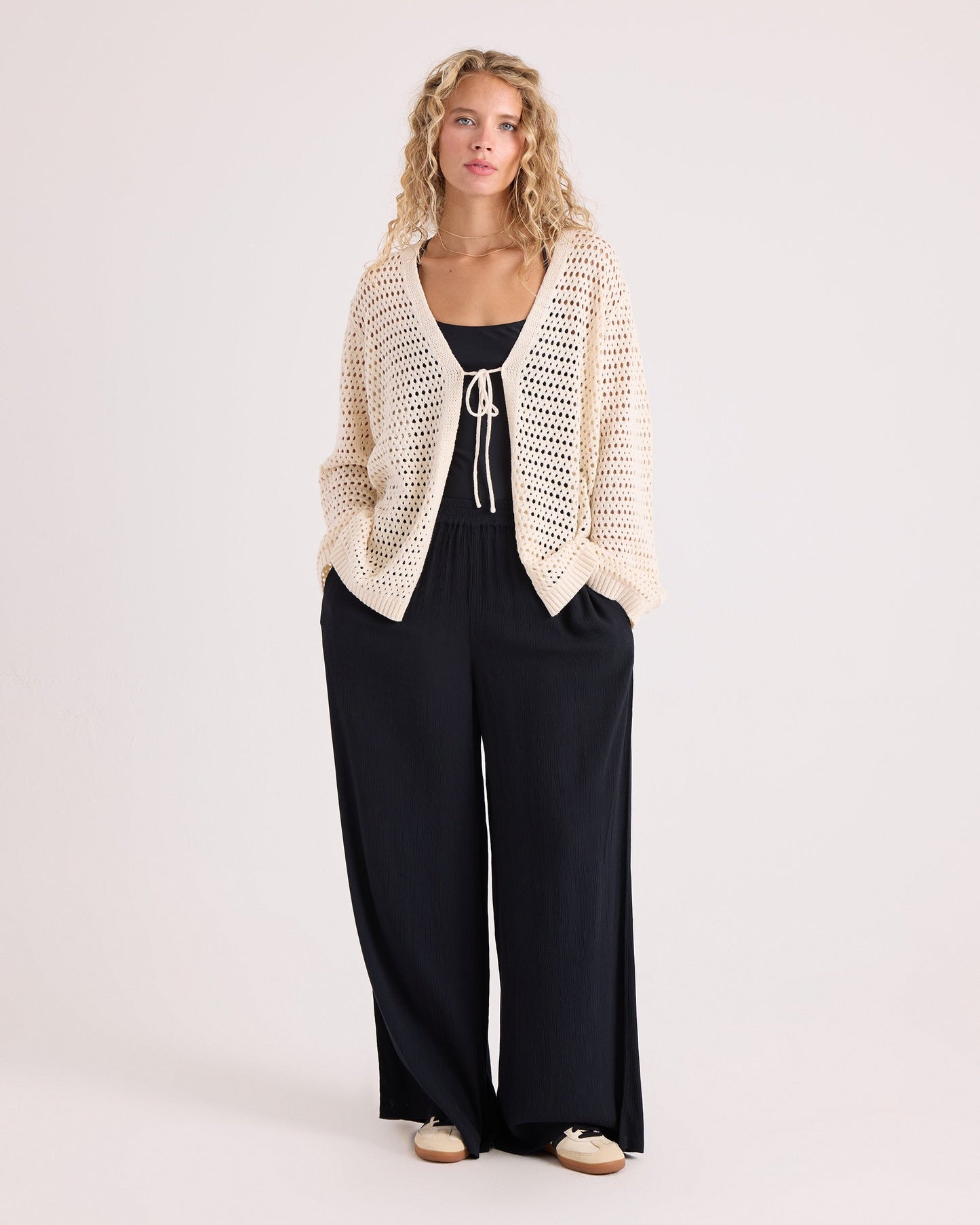 Wearing the Volcom Women's Go Go Wrap, a person with long curly hair stands against a plain light background with their hands in their pockets, layering the cream cotton-acrylic blend cardigan over a black top and loose black pants.