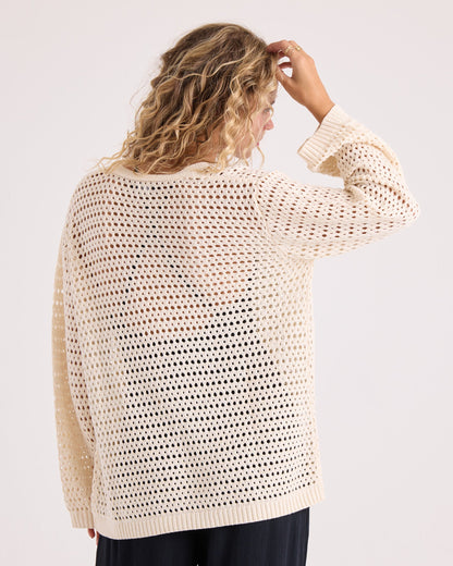 A person with wavy blonde hair stands with their back to the camera and hand raised to their head against a plain background, wearing the Volcom Women's Go Go Wrap—a loose, cream-colored, open-knit sweater top crafted from a soft cotton-acrylic blend—paired with black pants.