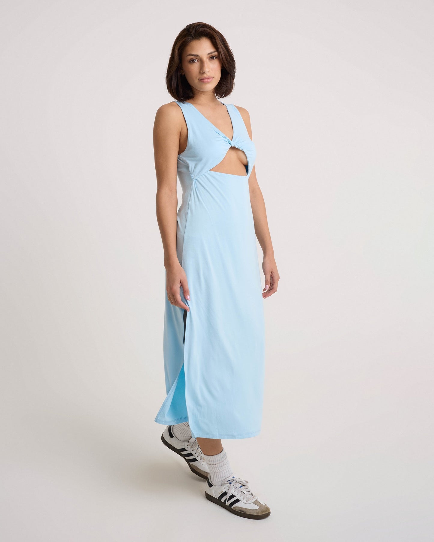 A woman in a Volcom Women's Stone Luz Dress with a twist-front bodice and front ring detail stands against a plain background. She pairs the sleeveless, cut-out midi dress with white socks and black-and-white sneakers.
