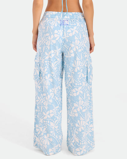 A person is seen from the back wearing Volcom Women's Wide Leg Pull On Pants in high-waisted light blue with a white floral and leaf print, soft cotton-viscose blend, side cargo pockets, elasticated waistband, and relaxed fit.