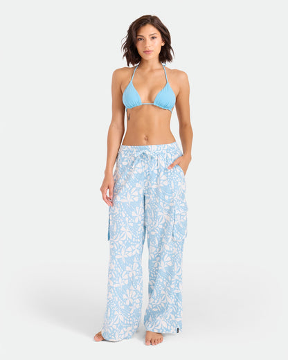 A woman stands barefoot against a white background, wearing a light blue bikini top and Volcom Women's Wide Leg Pull On Pants with a white floral pattern and elasticated waistband, her hands in her pockets.