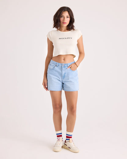 A woman stands against a plain background wearing the Volcom Women's Flip Side 1991 Mid Tee—a vintage-inspired, fitted white cropped shirt—paired with light blue denim shorts, white sneakers, and white socks featuring red and blue stripes.