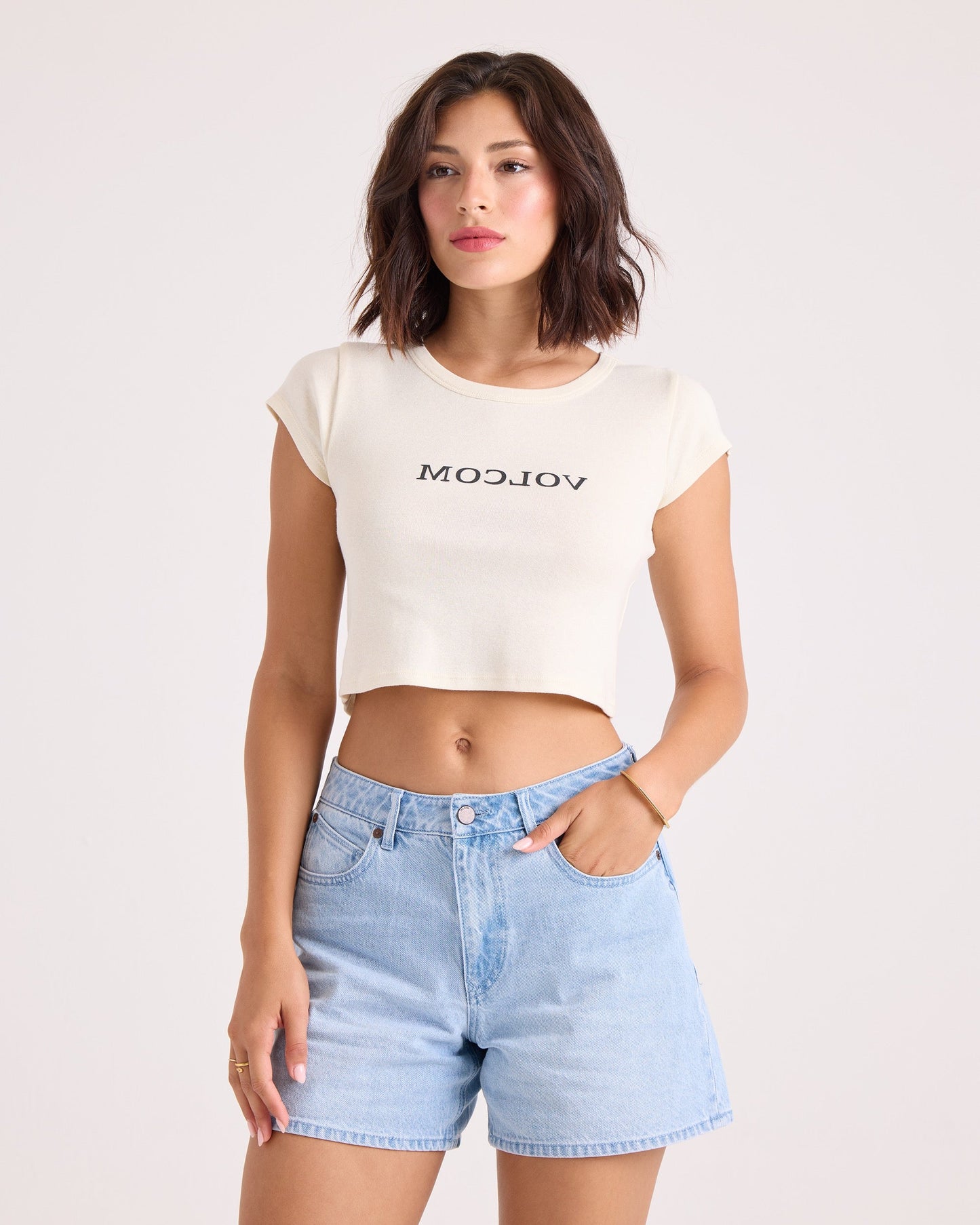 A woman with short brown hair wears the Volcom Women's Flip Side 1991 Mid Tee in vintage white, featuring a backward VOLCOM graphic, paired with light blue high-waisted denim shorts. She stands with one hand in her pocket against a plain light background.