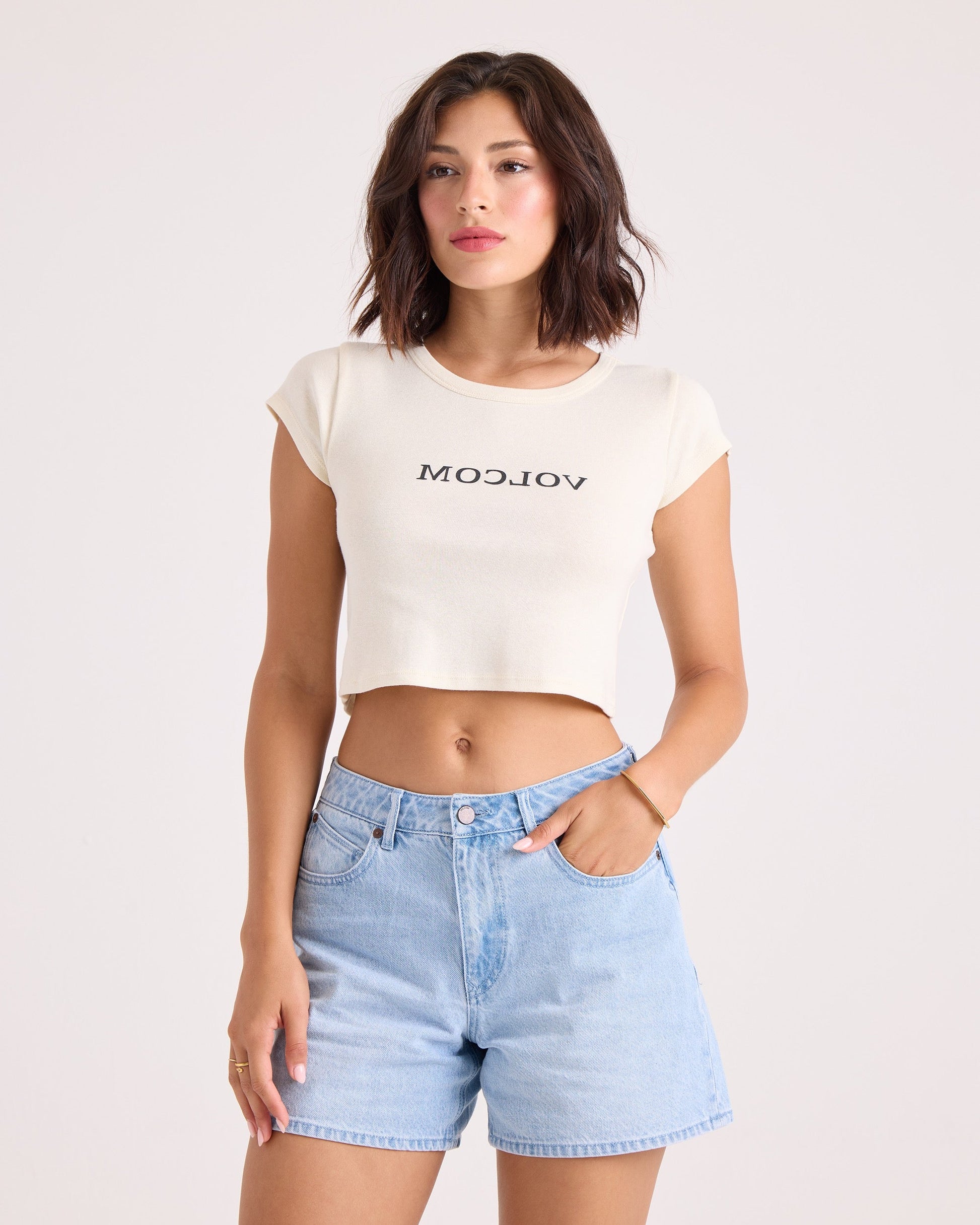A woman with short brown hair wears the Volcom Women's Flip Side 1991 Mid Tee in vintage white, featuring a backward VOLCOM graphic, paired with light blue high-waisted denim shorts. She stands with one hand in her pocket against a plain light background.