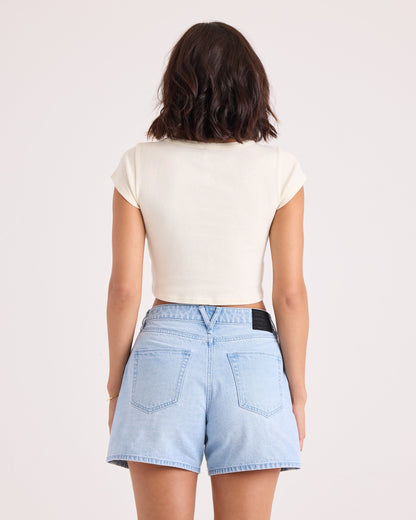 A woman with short dark hair is standing with her back to the camera, wearing the Volcom Women's Flip Side 1991 Mid Tee in vintage white and light blue denim shorts against a plain light background.