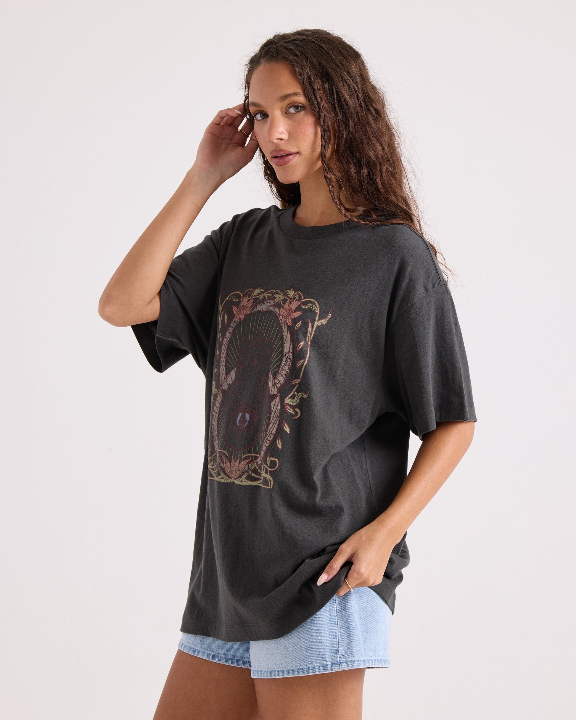 A woman with long wavy hair poses against a plain background, wearing the Volcom Women's Gatekeeper Stones Throw Tee—an oversized black vintage-style t-shirt made from 100% cotton—and light denim shorts, with one hand near her face and the other in her pocket.