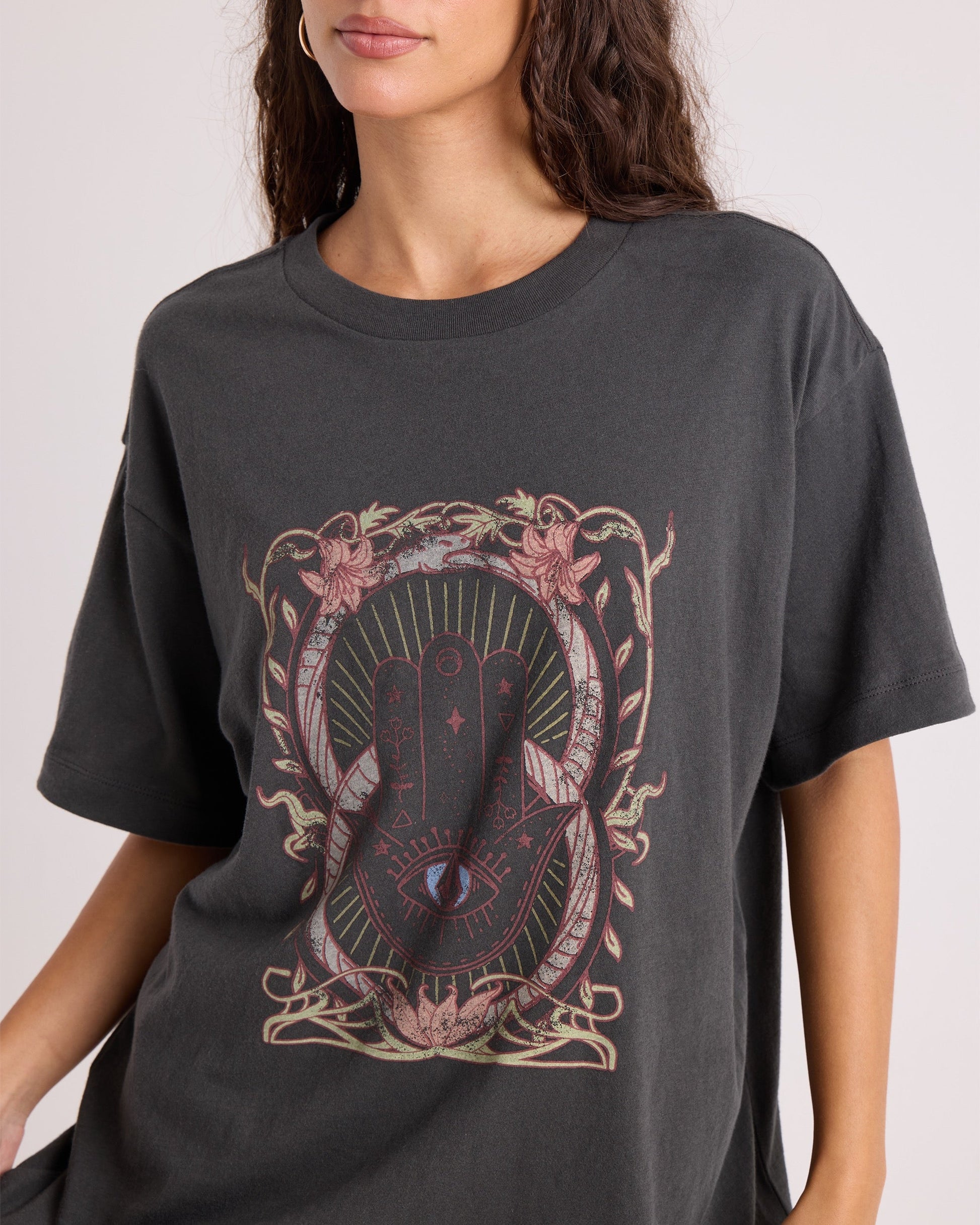 A woman wears the Volcom Women's Gatekeeper Stones Throw Tee—an oversized vintage-style black T-shirt made from 100% cotton, showcasing a mystical hand-with-eye motif at the center and surrounded by ornate floral and geometric patterns.