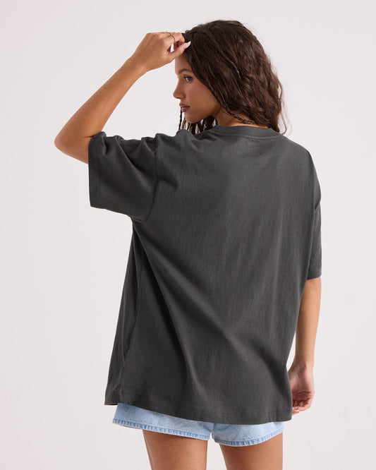 A woman with wavy brown hair stands with her back to the camera, touching her hair, wearing the Volcom Women's Gatekeeper Stones Throw Tee—an oversized vintage black t-shirt made from 100% cotton—paired with light denim shorts. The background is plain and light-colored.