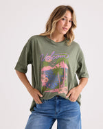 Wearing the Volcom Women's Night Shade Bad Tee—a relaxed, oversized green graphic shirt featuring a vibrant pink and blue tropical print—a woman stands against a plain background with one hand in her jean pocket, gazing confidently at the camera.