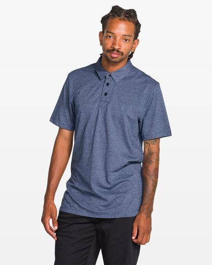 A man with dreadlocks and a tattooed left arm models the Volcom Men's Wowzer Polo Short Sleeve Shirt in blue, styled with black pants, standing against a plain white background.