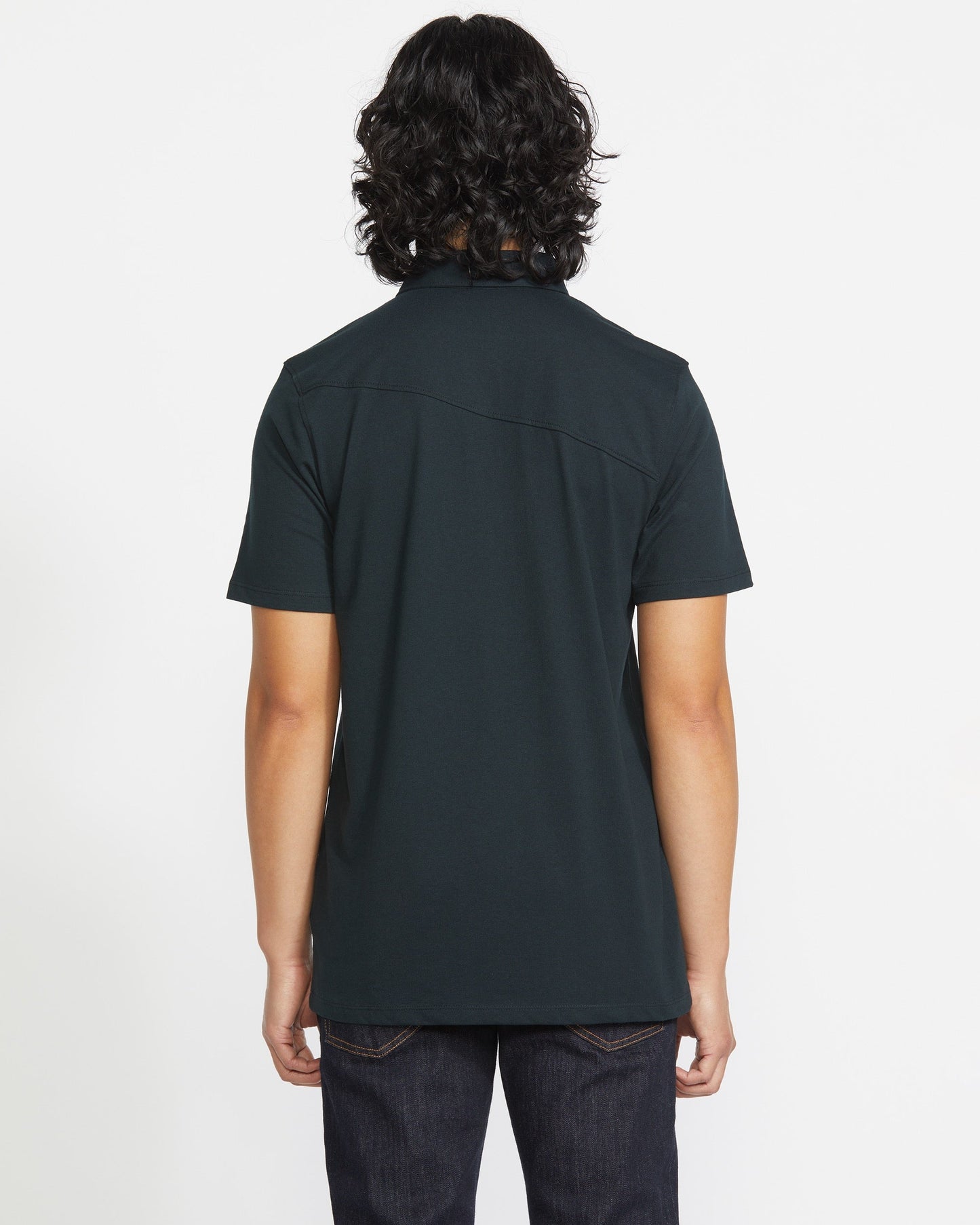 A person with shoulder-length wavy dark hair, wearing a Volcom Men's Wowzer Polo Short Sleeve Shirt in a dark cotton-poly blend and dark jeans, stands with their back to the camera against a plain white background.