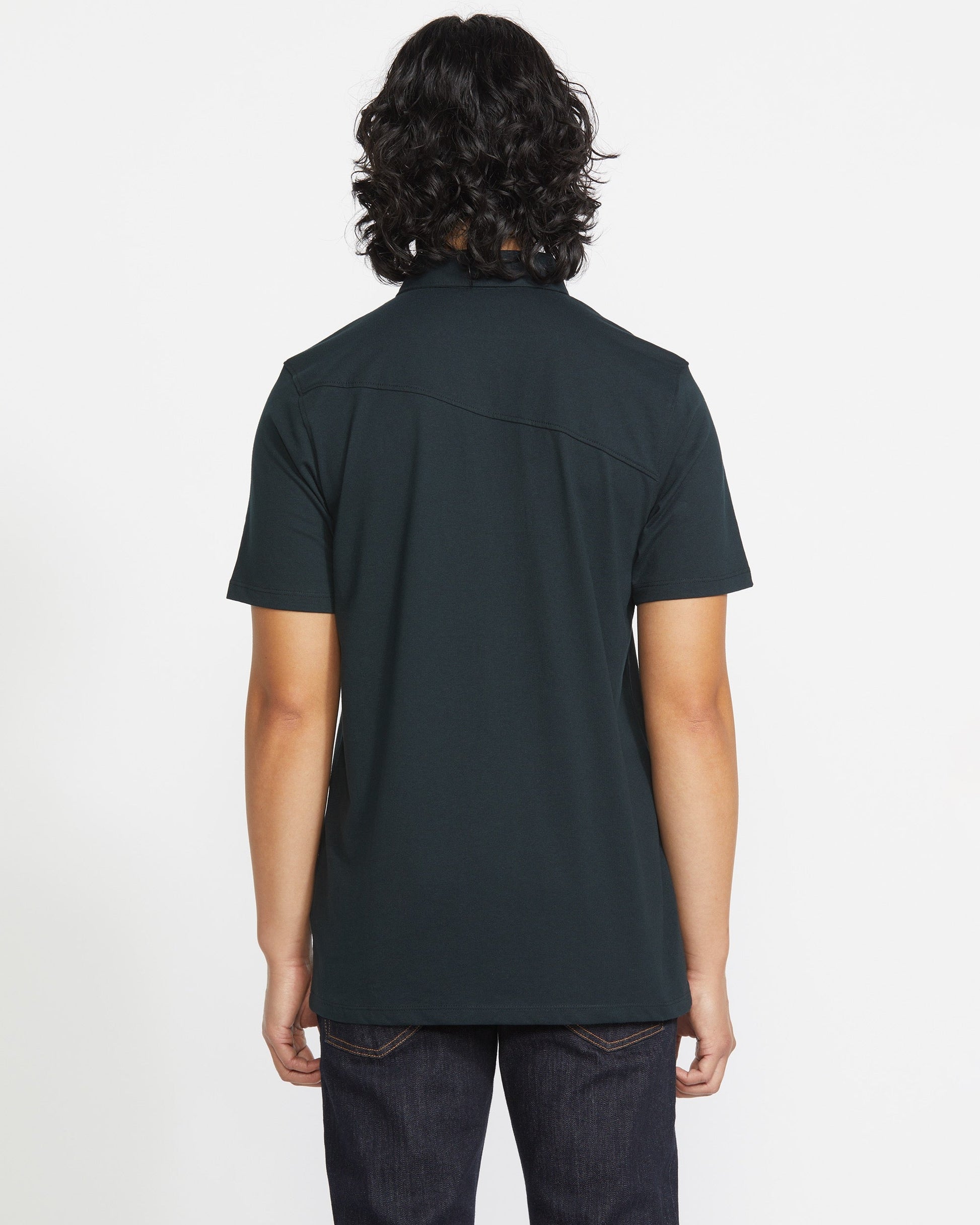 A person with shoulder-length wavy dark hair, wearing a Volcom Men's Wowzer Polo Short Sleeve Shirt in a dark cotton-poly blend and dark jeans, stands with their back to the camera against a plain white background.