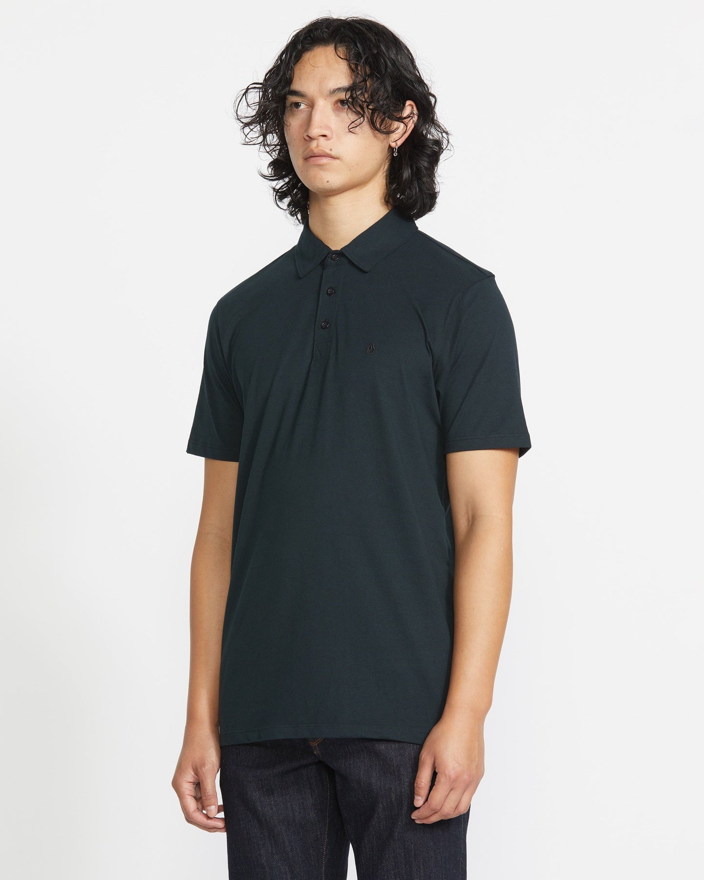 A man with long dark hair stands against a plain white background wearing the Volcom Men's Wowzer Polo Short Sleeve Shirt and dark pants, looking slightly to the side with a neutral expression.
