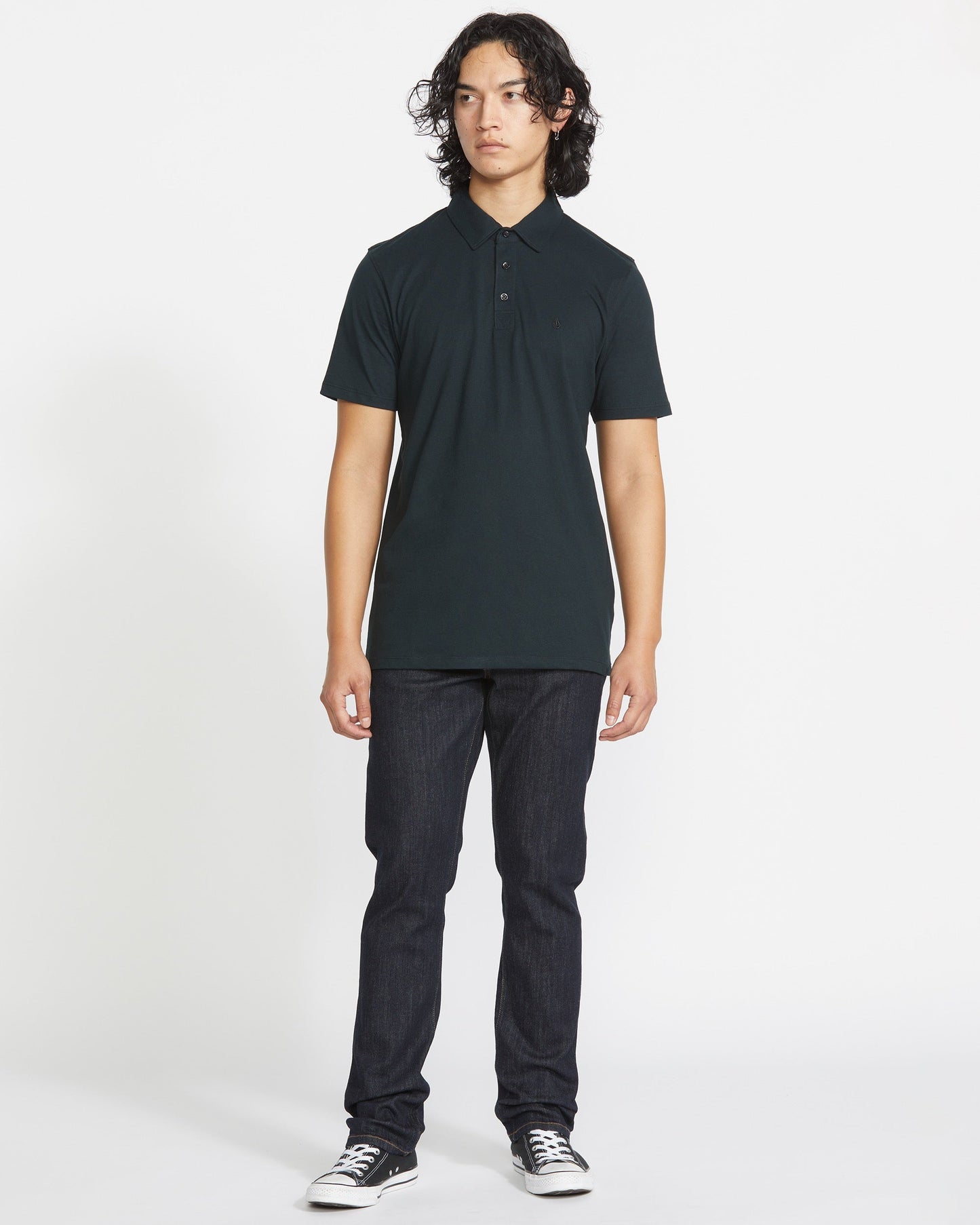 A person with long, curly dark hair stands facing forward in a Volcom Men's Wowzer Polo Short Sleeve Shirt, paired with dark jeans and black low-top sneakers against a plain white background.