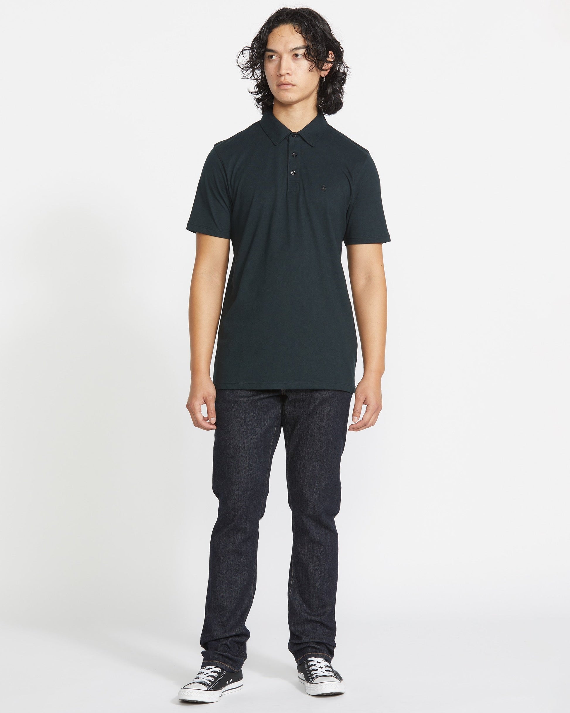 A person with long, curly dark hair stands facing forward in a Volcom Men's Wowzer Polo Short Sleeve Shirt, paired with dark jeans and black low-top sneakers against a plain white background.
